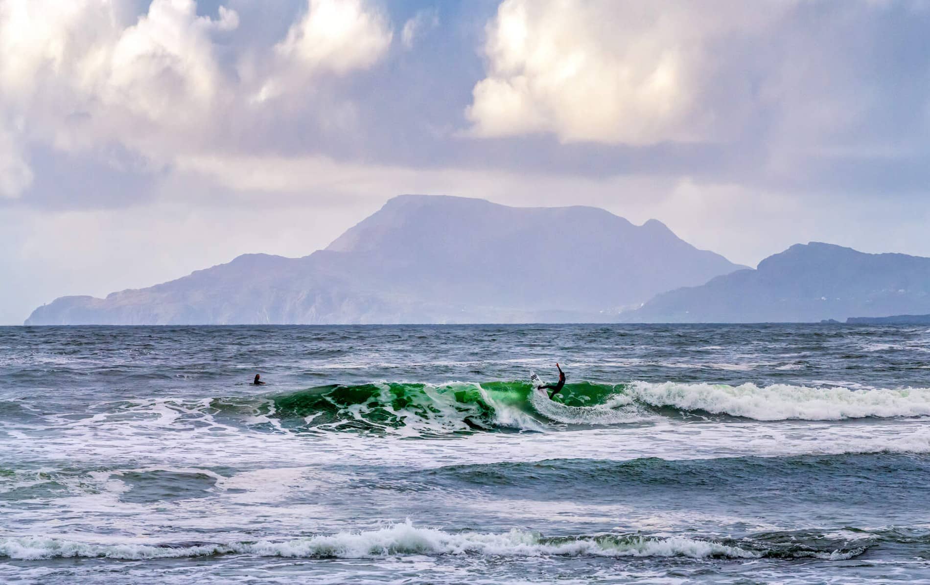 Surfista montando una ola en Tullan Strand, Bundoran, con montañas brumosas al fondo y un espectacular oleaje del Atlántico.