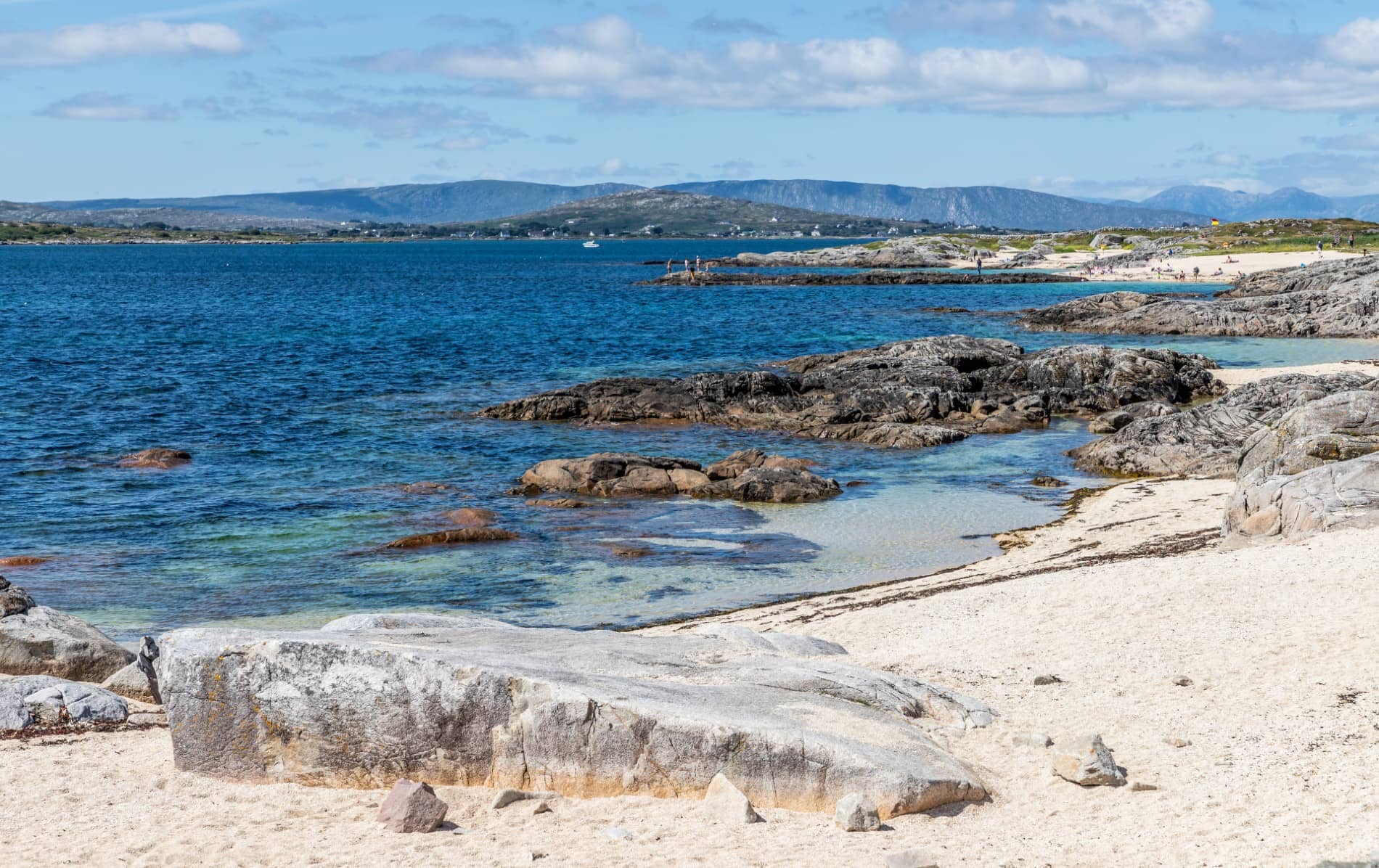 Aguas azules cristalinas y costa de arena blanca en la playa de Carraroe, Galway, con afloramientos rocosos y colinas más allá.