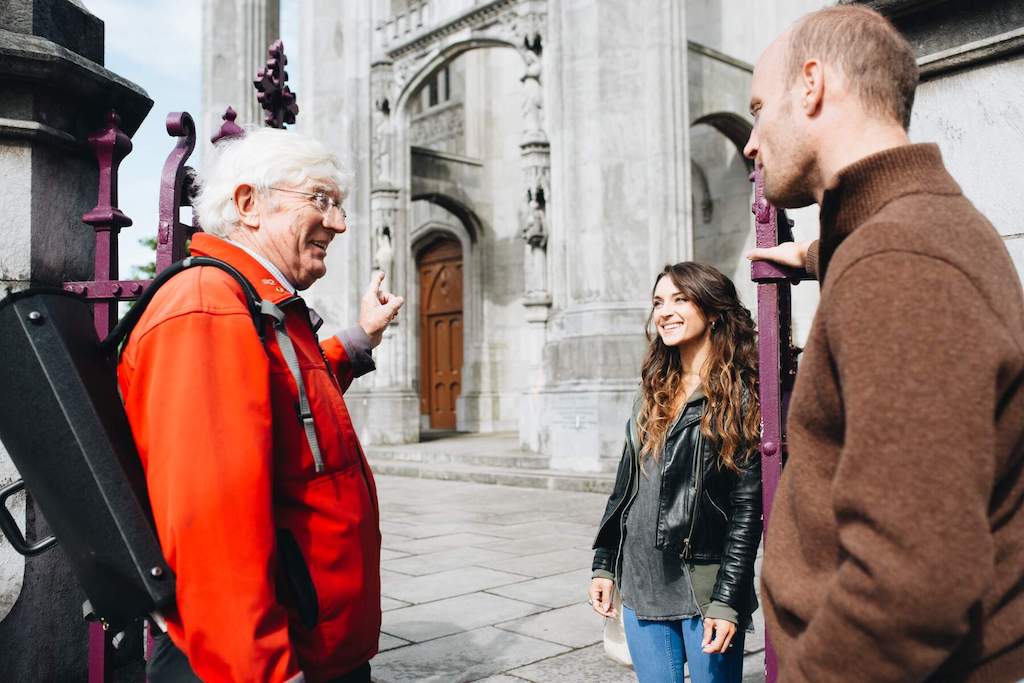 Guide touristique en veste rouge faisant des gestes tout en parlant à deux visiteurs souriants devant une cathédrale dans la ville de Cork.