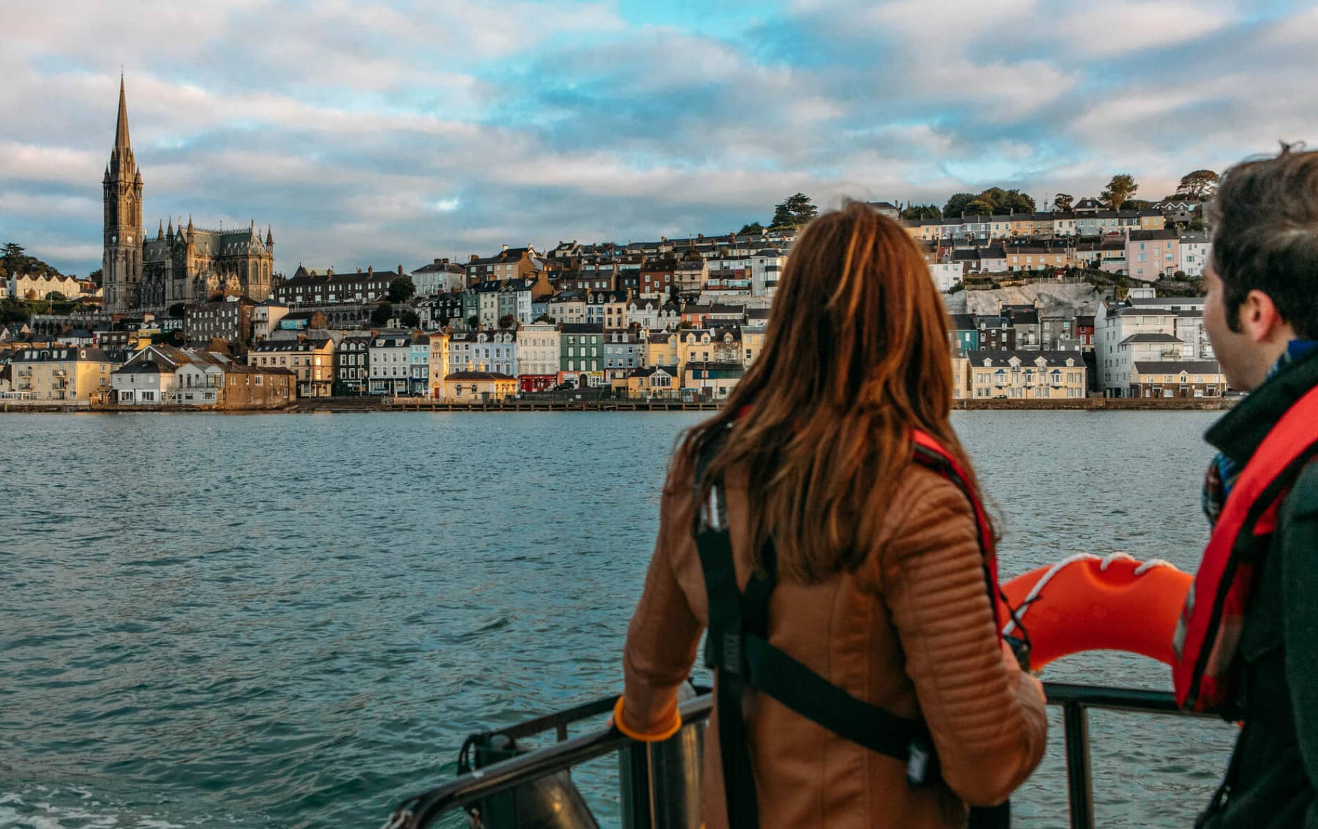 Deux passagers du ferry en gilets de sauvetage regardent les bâtiments pastel et la cathédrale St Colman qui s'élèvent au-dessus du front de mer de Cobh.