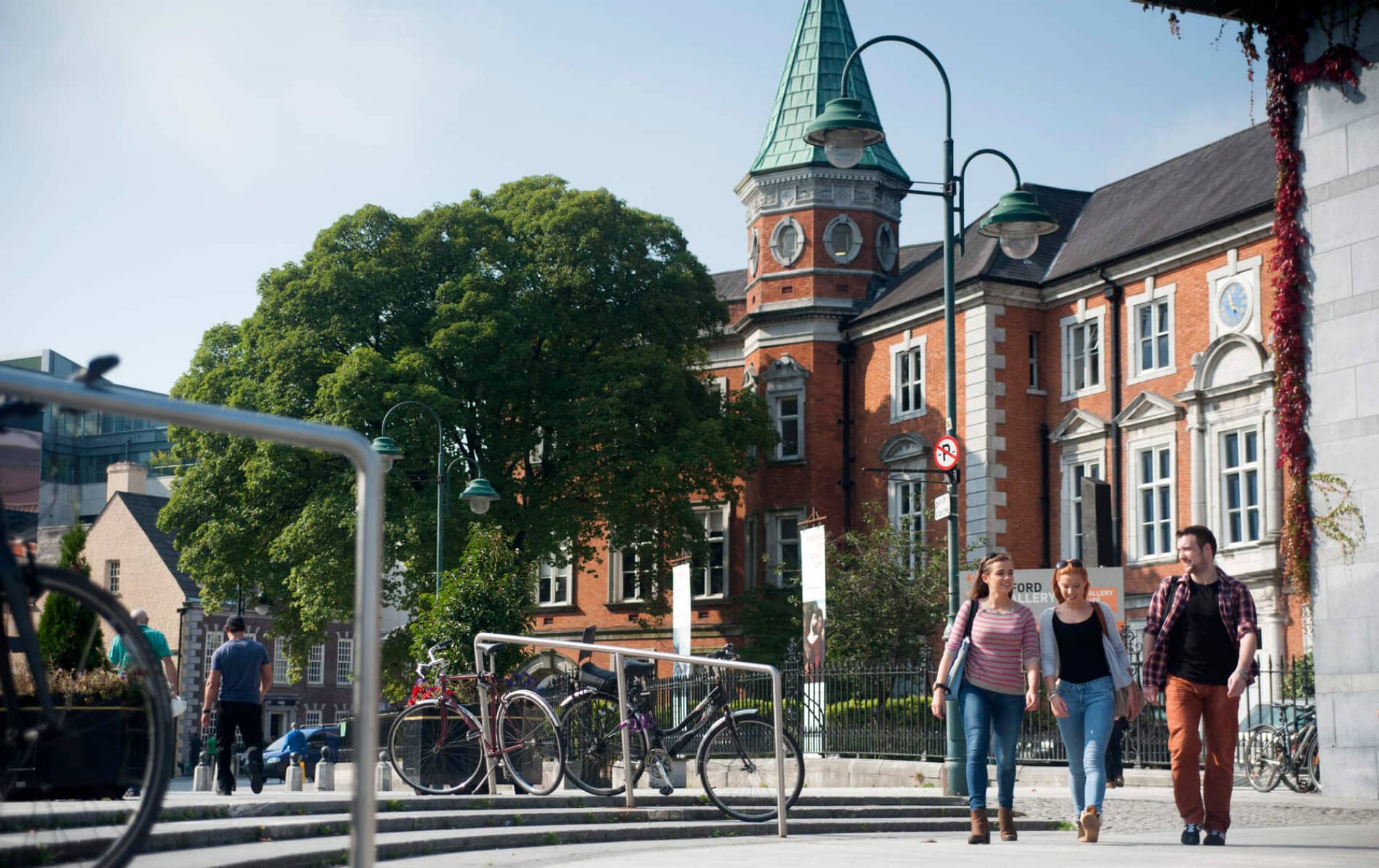 Trois personnes marchent près de la Crawford Art Gallery, un bâtiment en briques rouges avec une tour horloge, au centre-Ville de Cork.