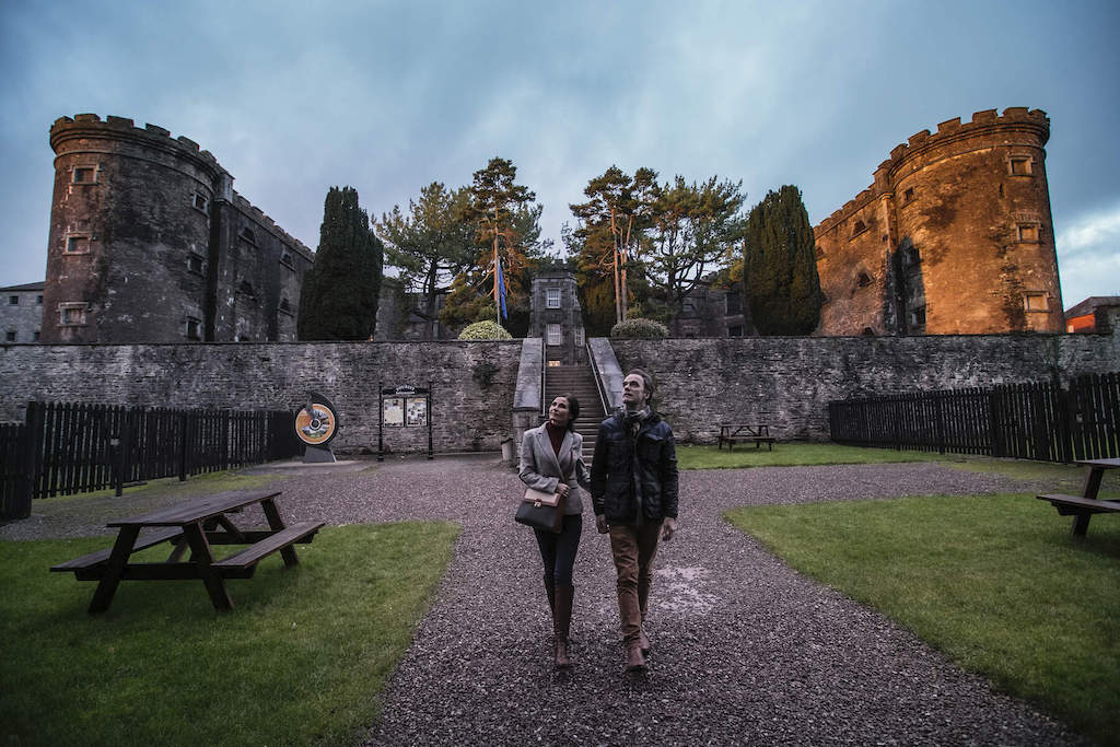 Un couple se promenant dans la cour de la prison de la ville de Cork, entouré de tours en pierre et d'arbres au crépuscule.