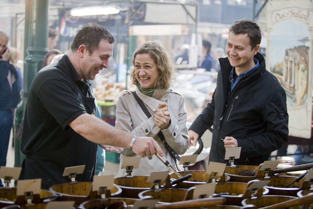 Un commerciante sorride mentre offre campioni a visitatori che ridono al St George’s Market, la storica sala da pranzo di Belfast.