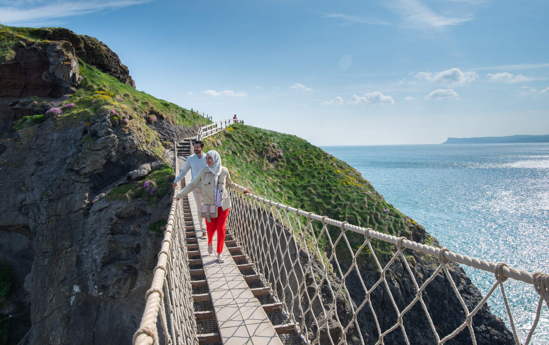 Koppel dat de Carrick-a-Rede hangbrug in County Antrim oversteekt, die tussen zeekliffen hangt met de oceaan eronder.