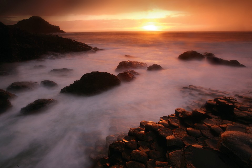 Les colonnes de basalte et les roches sombres rencontrent les vagues brumeuses à la chaussée des Géants, qui brillent d'orange sous un coucher de soleil éclatant.