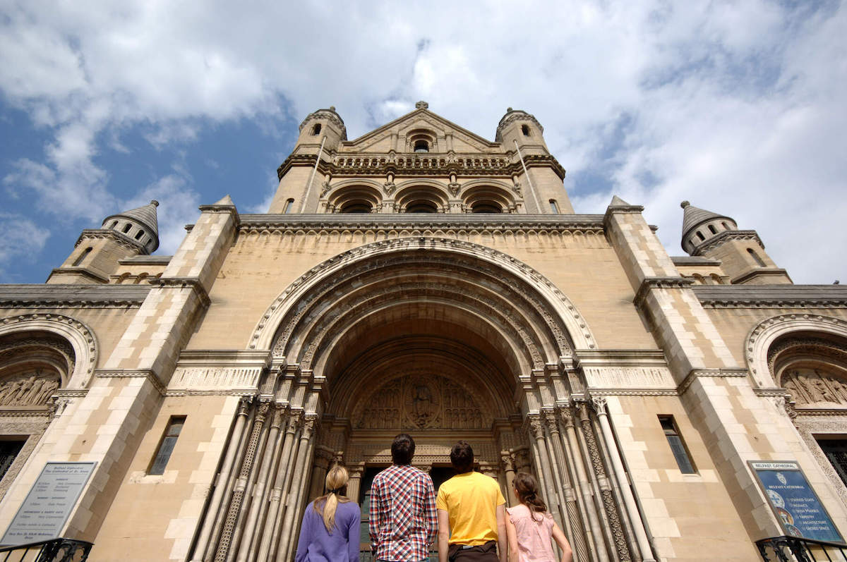 I visitatori guardano verso l'alto all'imponente ingresso romanico della Cattedrale di St Anne a Belfast in una giornata parzialmente nuvolosa.