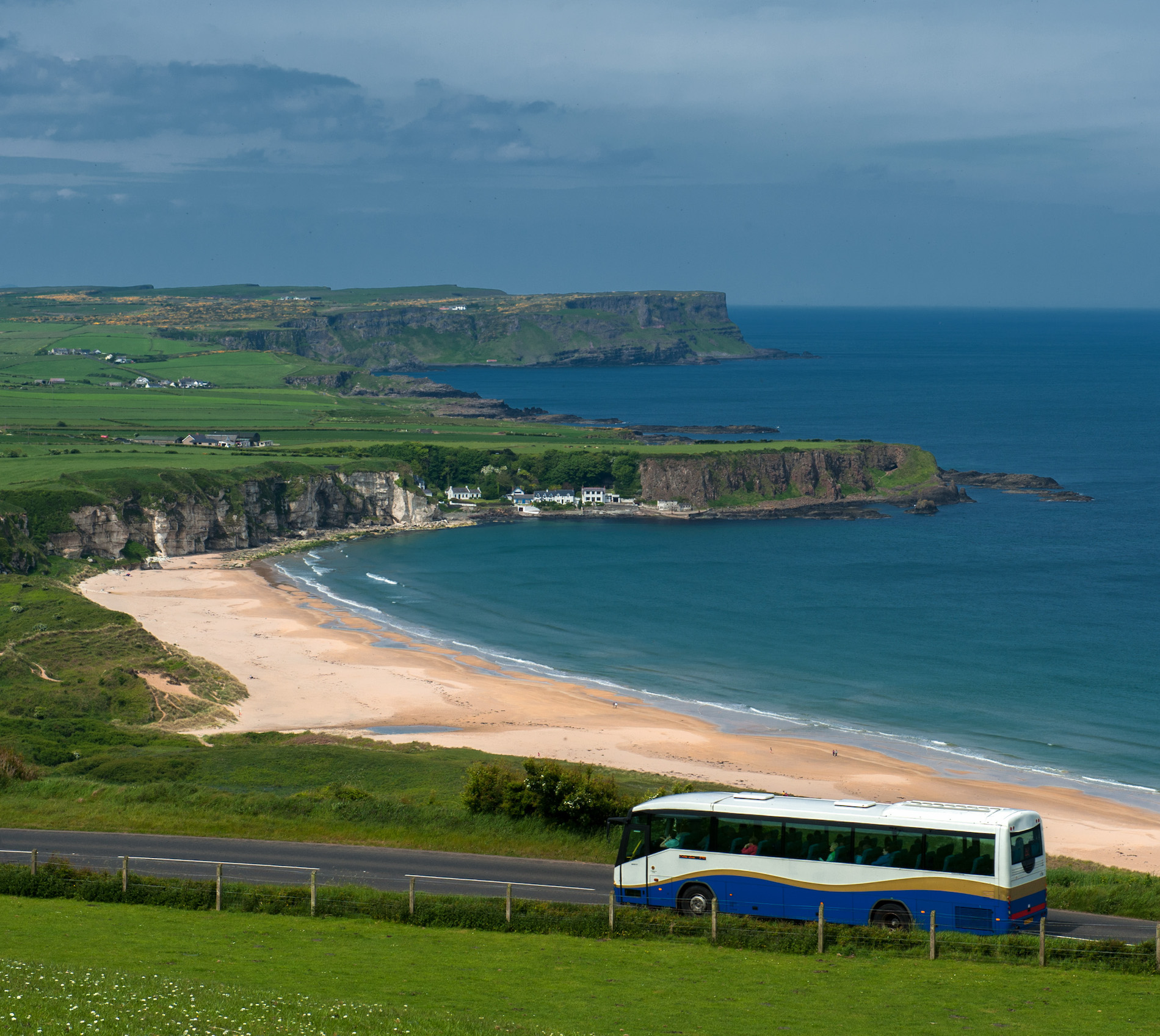 Coach driving past dramatic sea cliffs and a golden beach along Northern Ireland’s scenic Antrim coast.