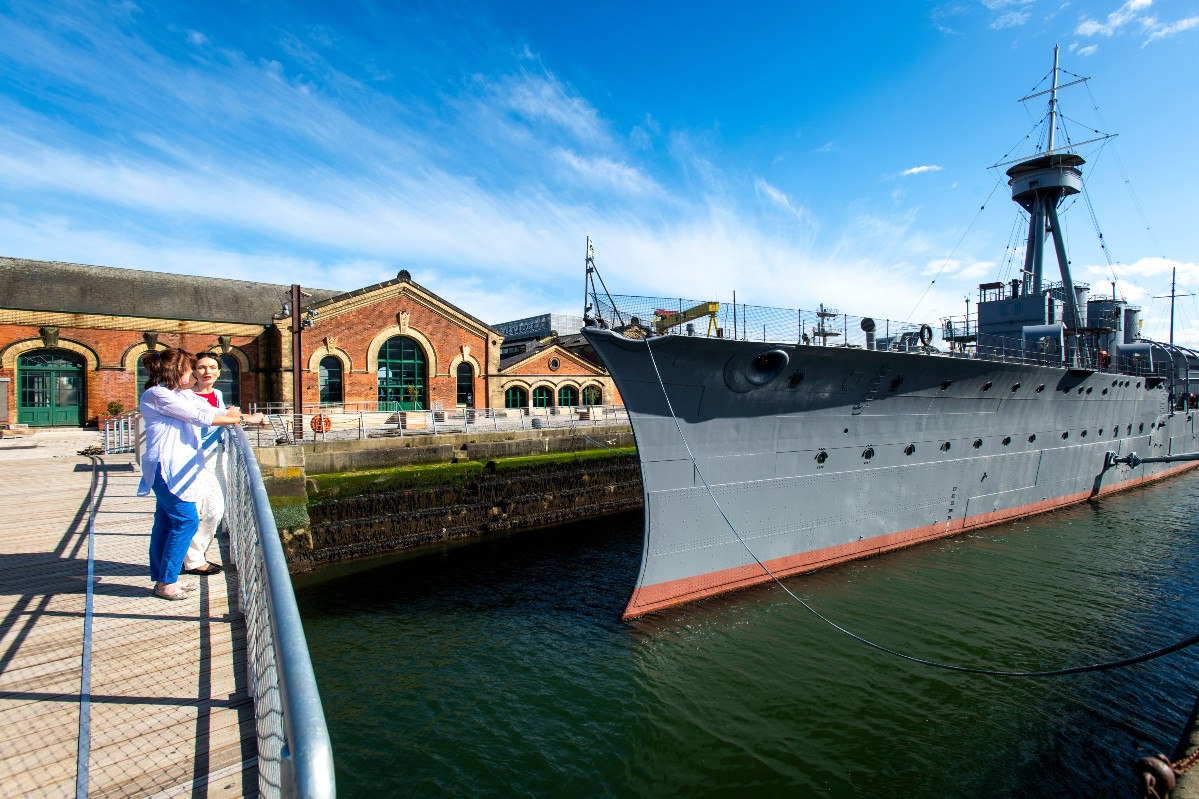 Due visitatori guardano l'HMS Caroline ormeggiata accanto ai magazzini in mattoni restaurati nel Titanic Quarter di Belfast.