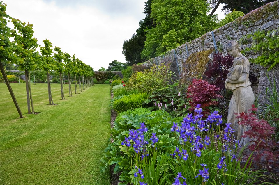 Jardins du château de Glenarm avec bordures soignées, statue en pierre d’une femme et d’un enfant, et des iris violets en fleurs.