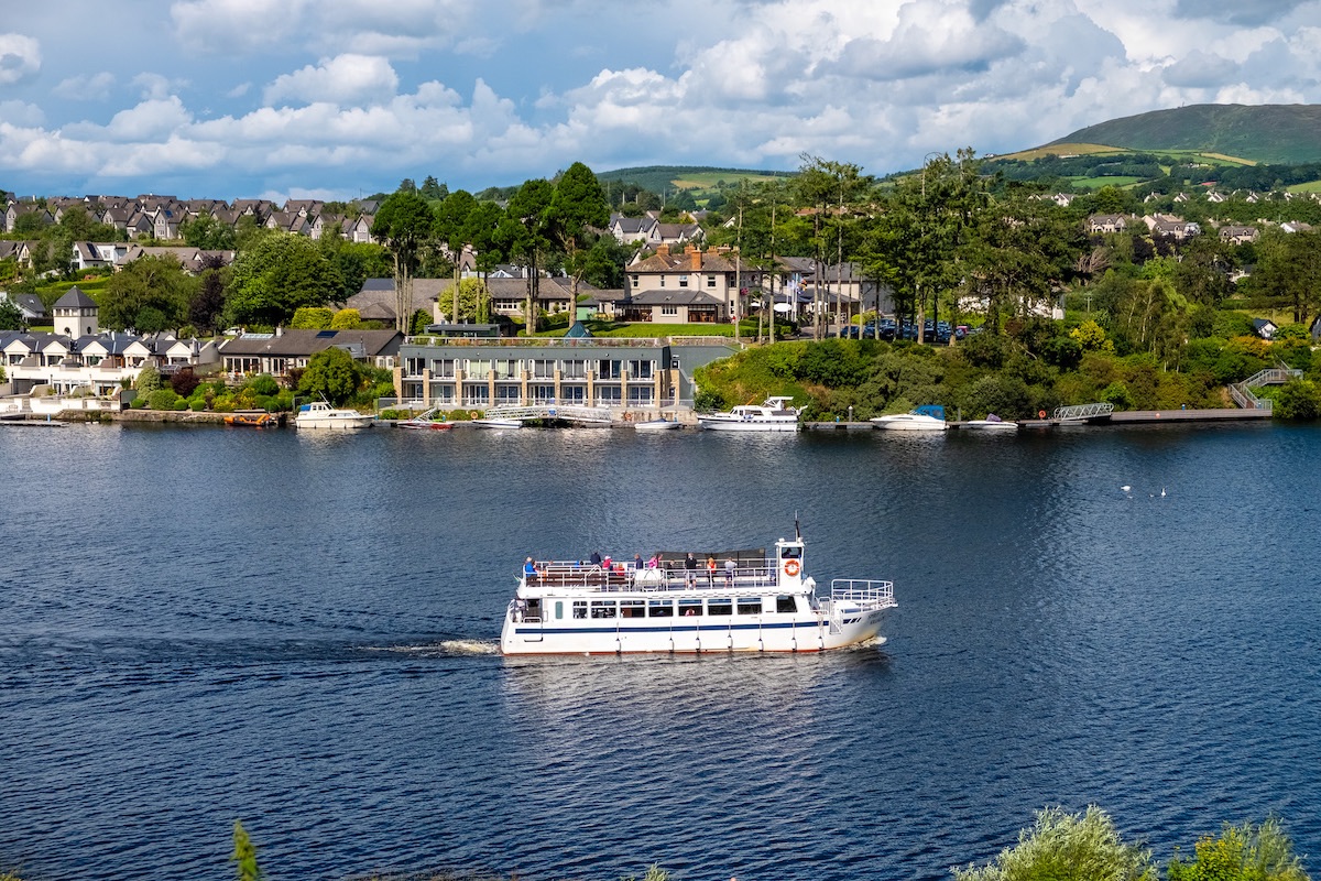 Weißer Flusskreuzer mit Passagieren, die an den blätterreichen Häusern am Ufer des Lough Derg, Clare, vorbeifahren.
