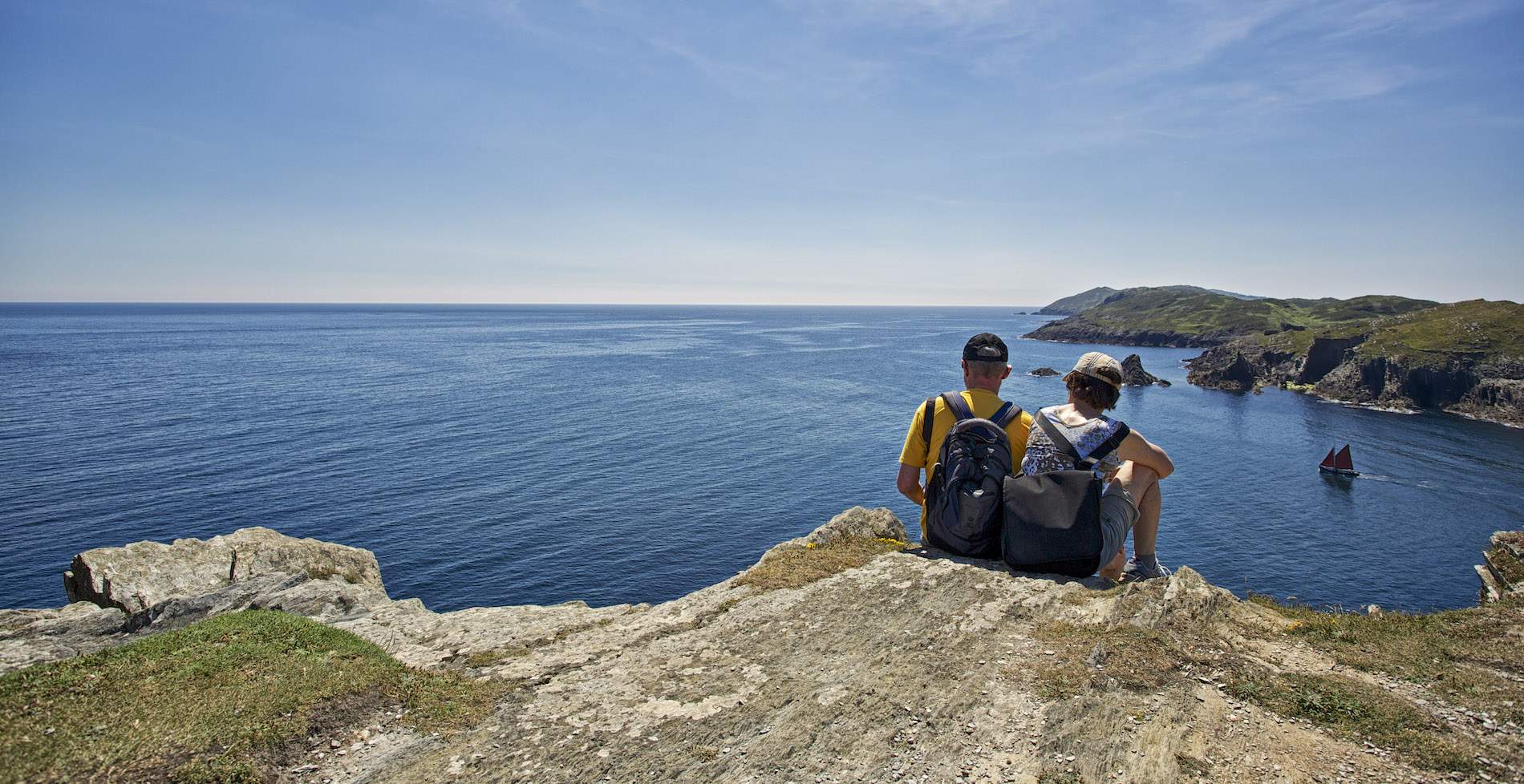 Two hikers sit on cliffs on the coast of County Kerry, overlooking the Atlantic Ocean as a sailboat passes below.
