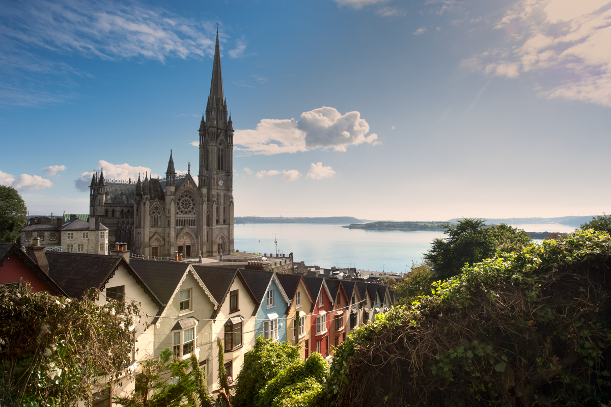 Row of brightly painted houses beneath the spire of St Colman’s Cathedral in Cobh, with Cork Harbour in the distance.