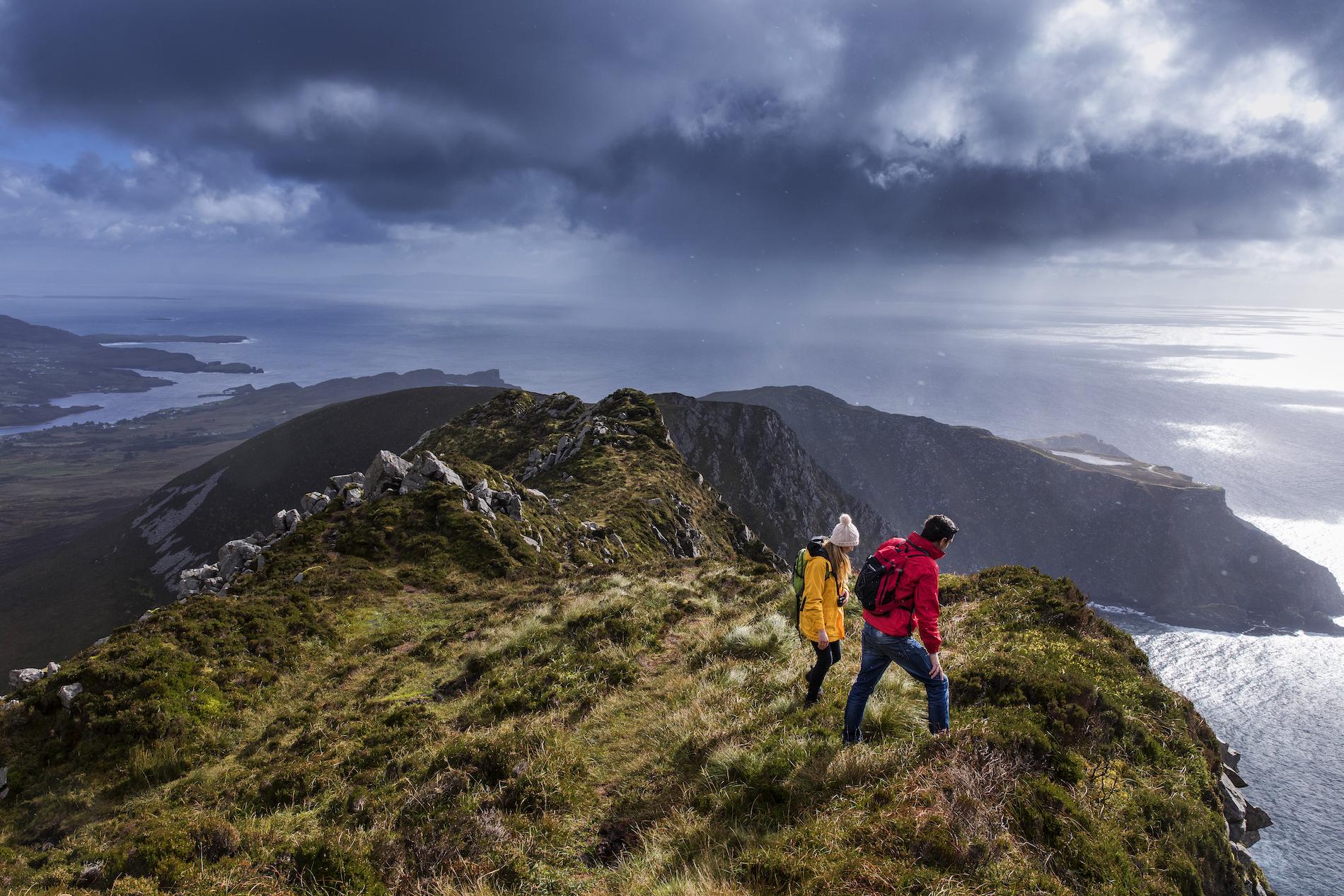 Walkers in bright jackets hiking along a high coastal ridge overlooking sea under heavy clouds.