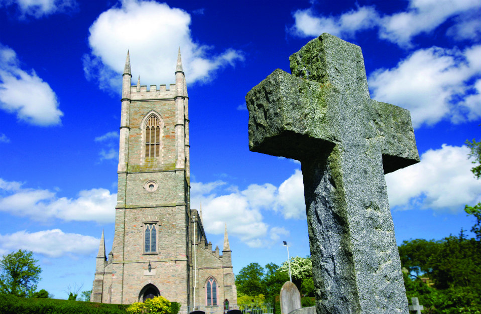 Down Cathedral, Downpatrick, met een stenen Keltisch kruis op de voorgrond op een heldere dag.
