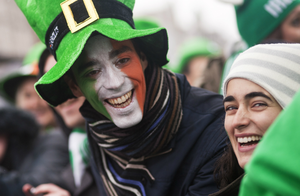 Twee mensen lachen, één met een gezicht geschminkt met de Ierse vlag en een groene hoed, en vieren de St. Patrick's Day in Ierland.