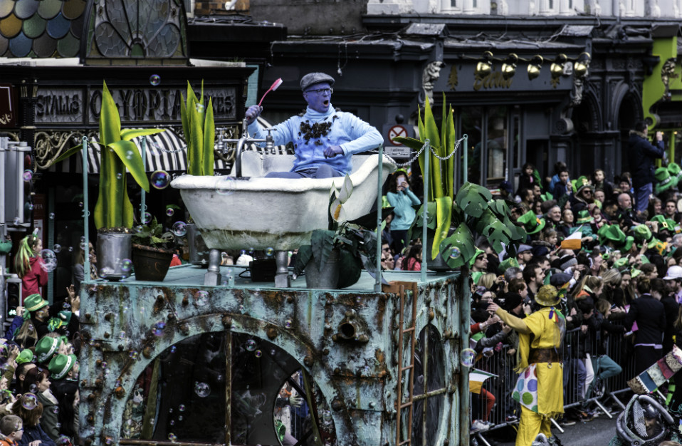 Een artiest met een blauwe schmink zit in een badkuip die als drijvende wagen wordt gebruikt en zwaait naar het publiek tijdens een St. Patrick's Day-parade in Ierland.