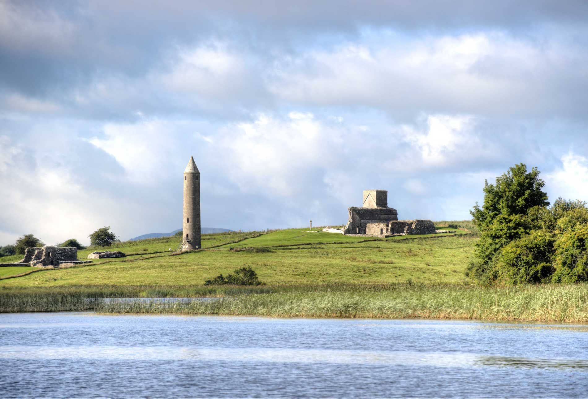 Medieval round tower and ruins on lush green hill beside lake under cloudy skies.