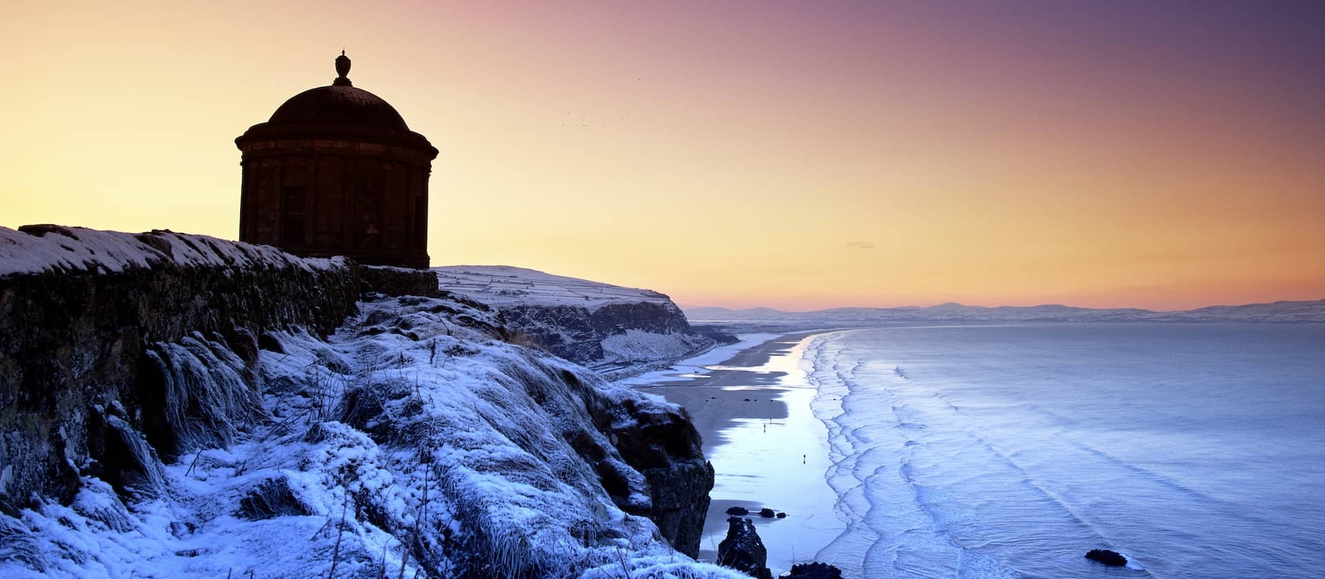 Mussenden Temple overlooks a snow-dusted coastline at sunset above Downhill Demesne in County Londonderry.