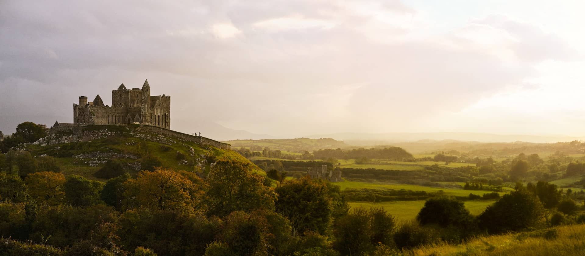 Soft golden light over the Rock of Cashel rising above rolling green and autumn-tinged fields under a hazy sky.