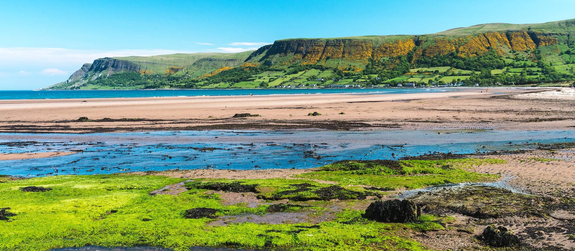 Waterfoot Beach in County Antrim met gouden zandstranden, kristalhelder water en groene heuvels onder een heldere lentehemel.