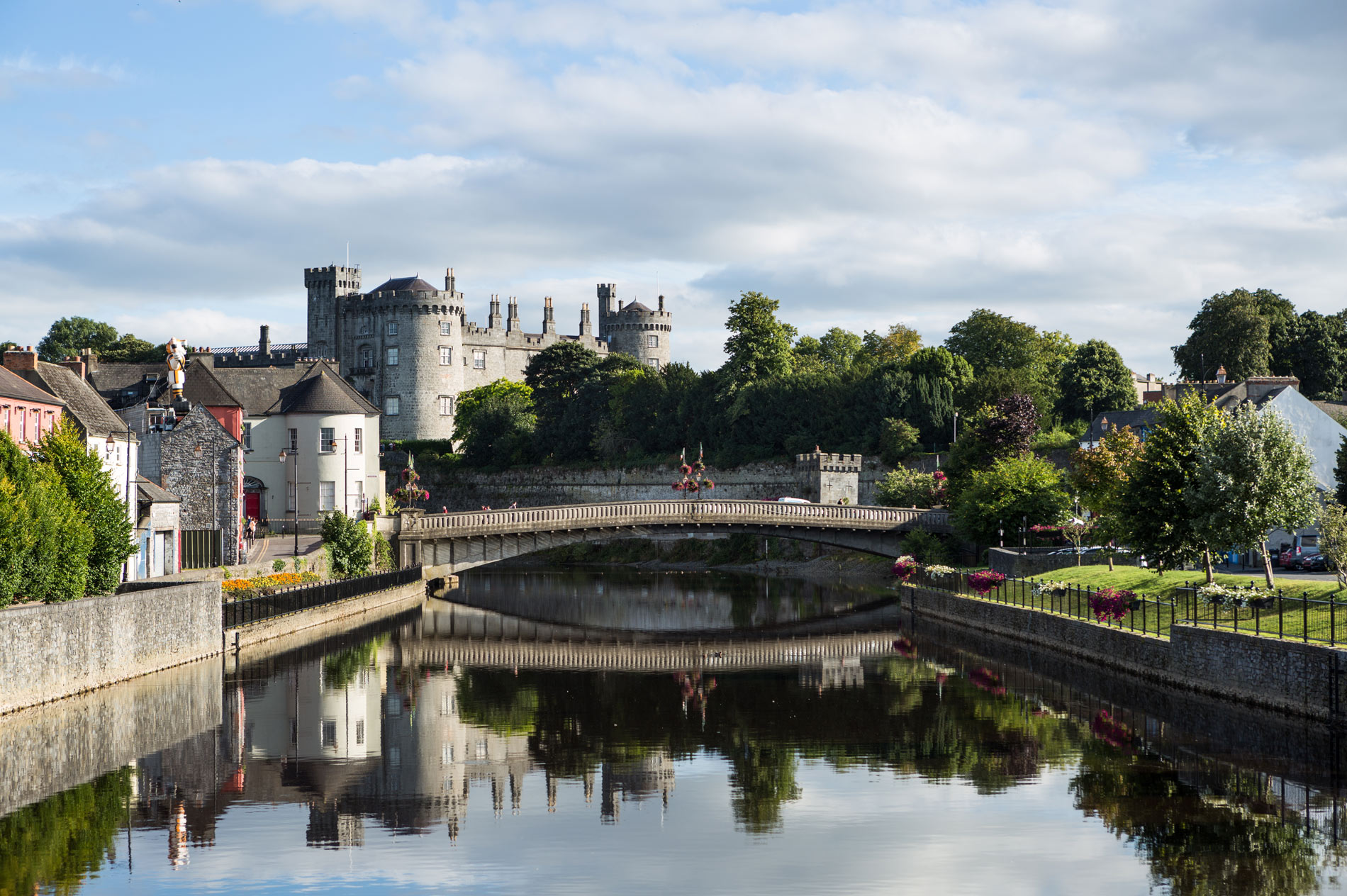 Stenen brug over de rivier de Nore met uitzicht op het kasteel van Kilkenny en oude stadsgebouwen, omgeven door bomen en bloemen.