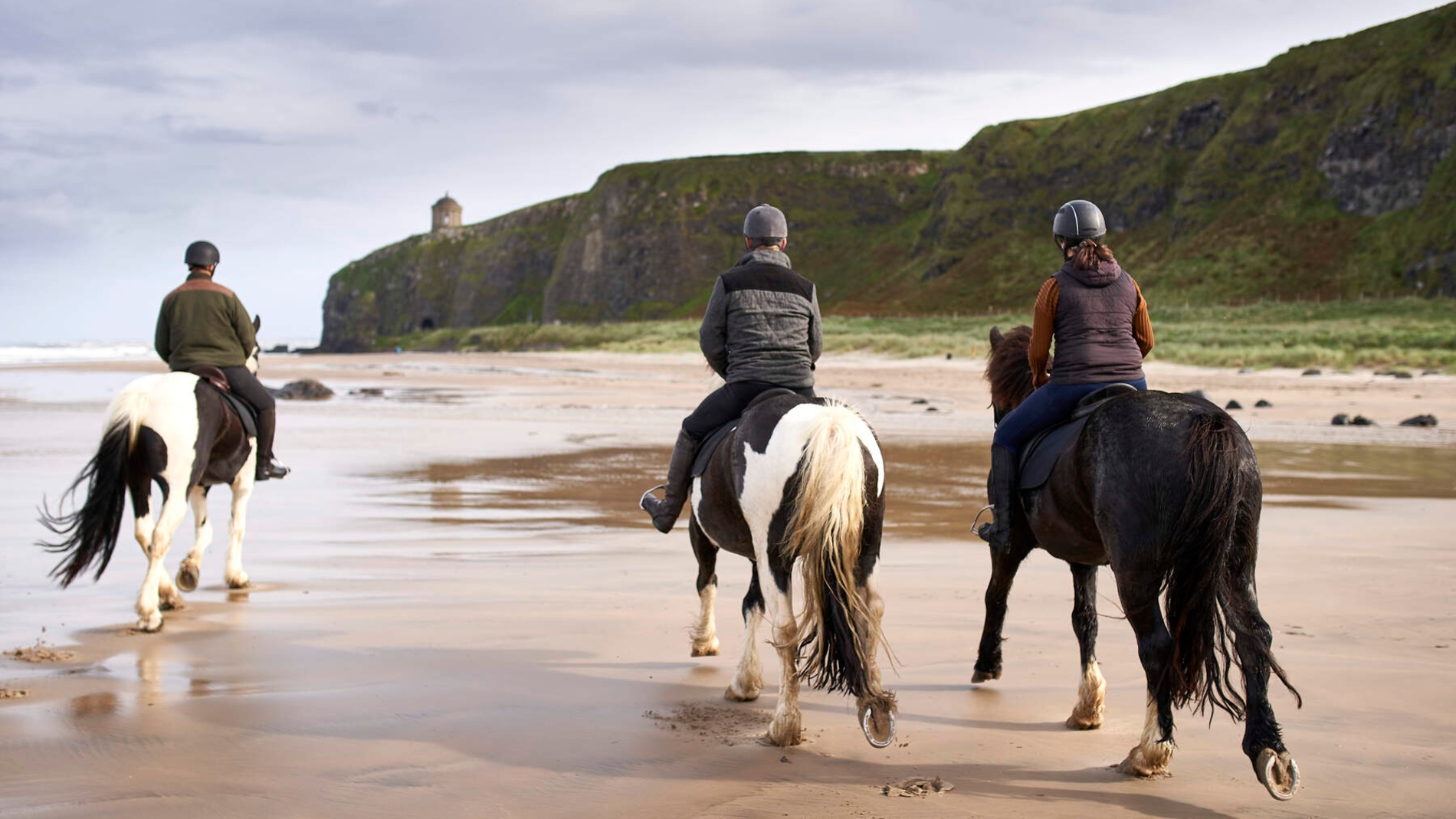 Mensen rijden te paard langs een zandstrand onder de Mussenden Tempel en kliffen langs de Causeway kust van Noord-Ierland.