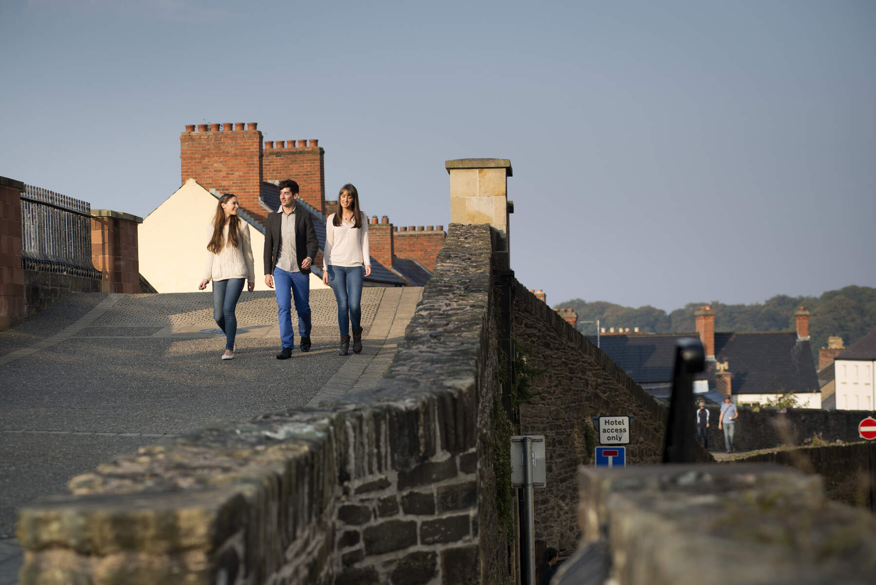 Drie vrienden lopen langs de 17th-century muren van Derry~Londonderry, met rode bakstenen schoorstenen en daken op de achtergrond.