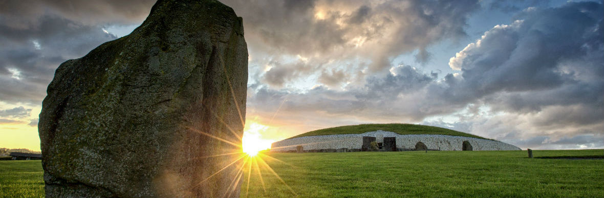 Lage winterzon die schittert naast een staande steen bij Brú na Bóinne, met de Newgrange-grafheuvel erachter.