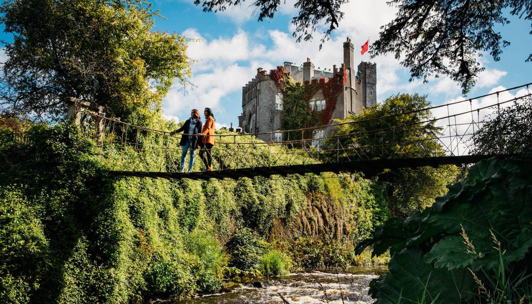 Deux personnes traversant un pont en corde avec le château de Birr, comté d'Offaly, en arrière-plan, entouré de verdure.