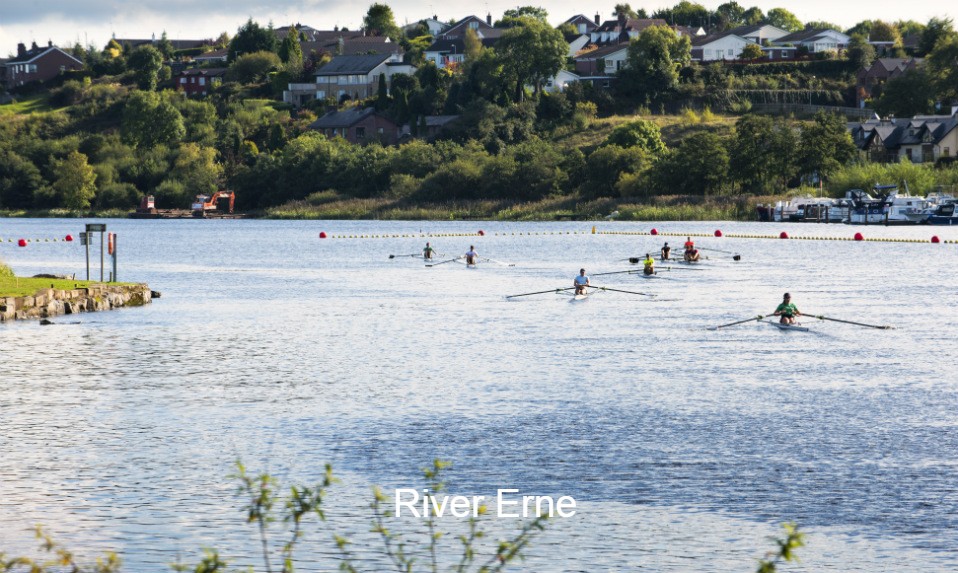 Rowers glide across the River Erne with lush greenery and riverside houses in the background.