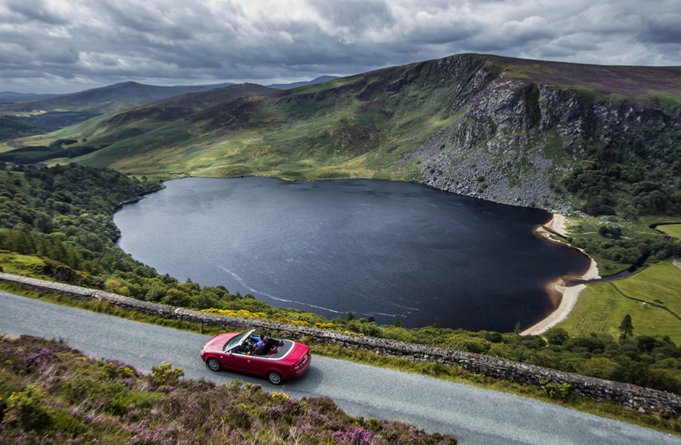 Red convertible driving past Lough Tay in Wicklow Mountains with dramatic valley scenery.