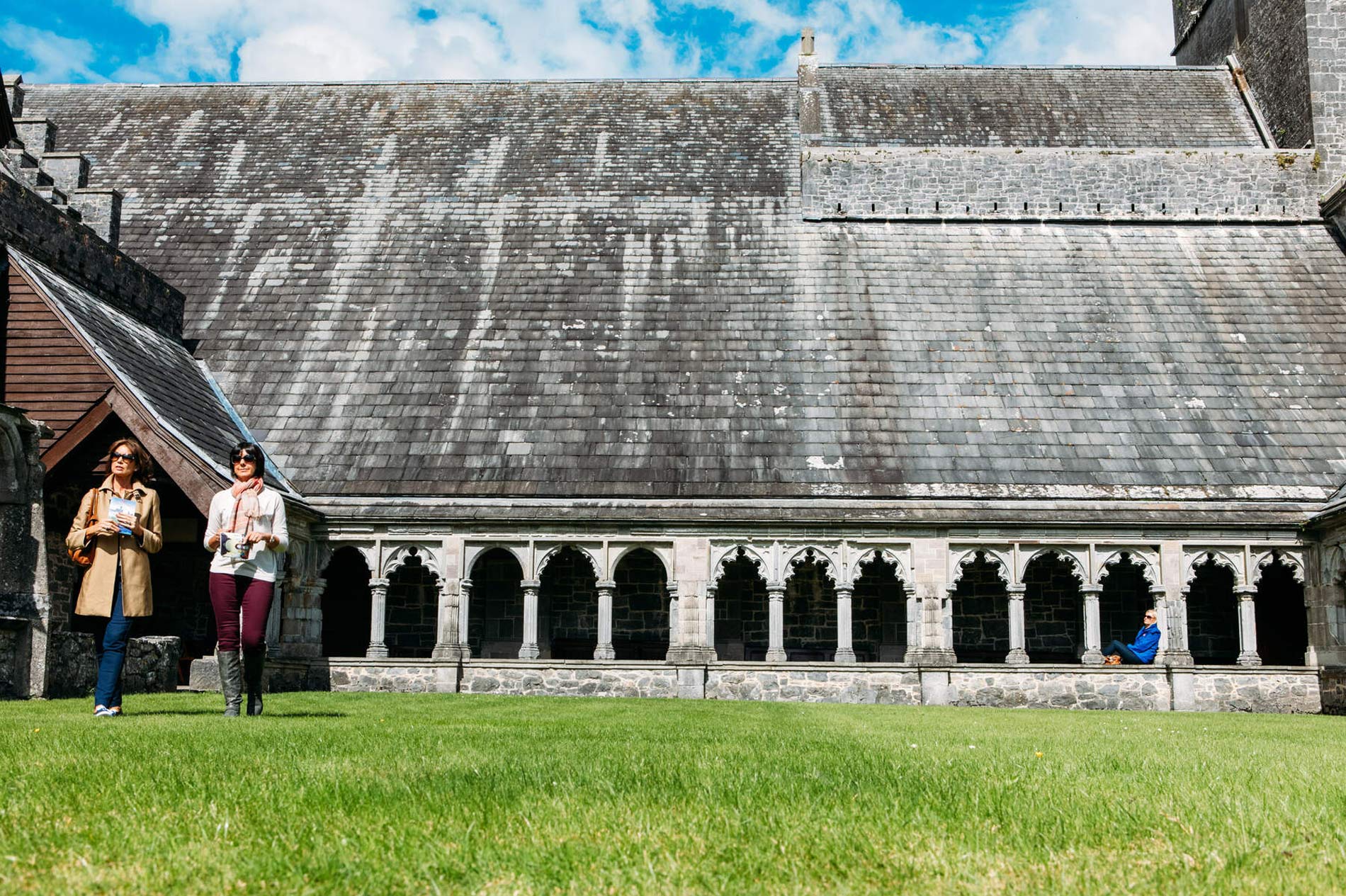 Deux femmes marchant devant les cloîtres d'une abbaye historique avec des arches en pierre et un toit en ardoise.