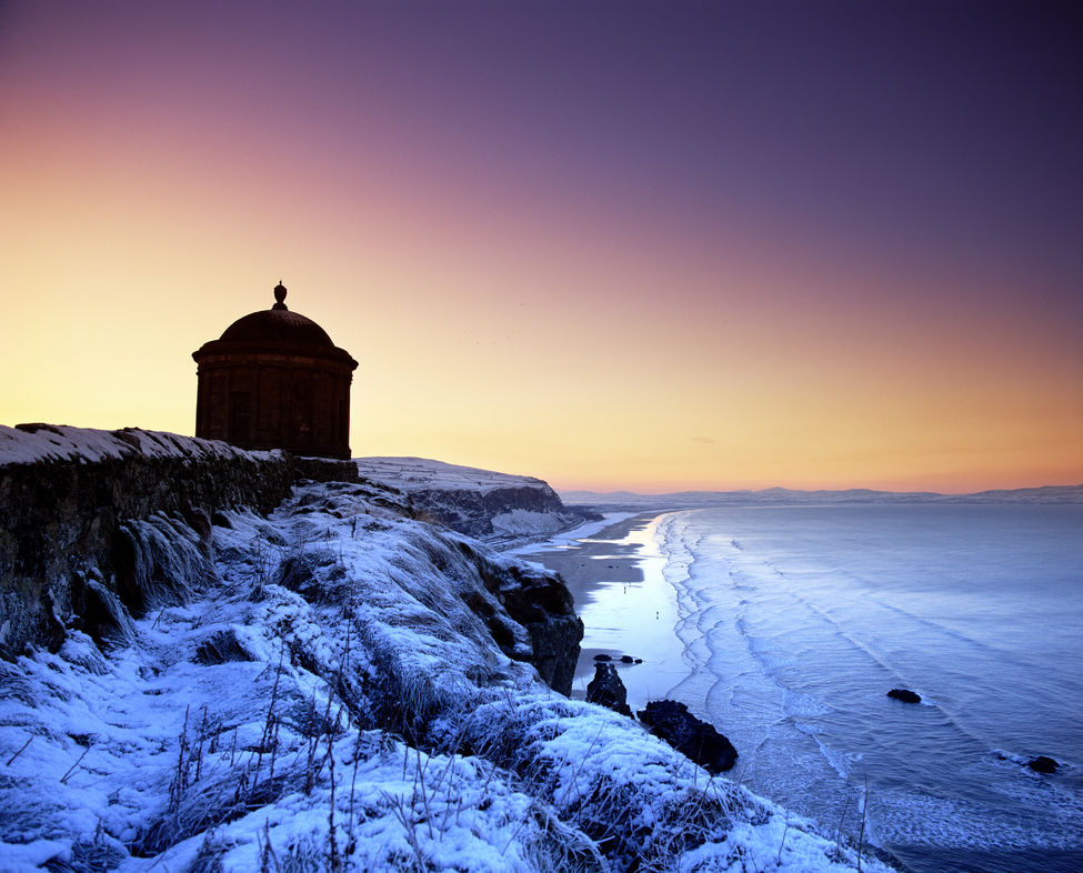 Schneigedeckte Klippen und der Mussenden-Tempel blicken auf den eisigen Strand und den sanften Wintersonnenuntergang in der Nähe von Castlerock herab.