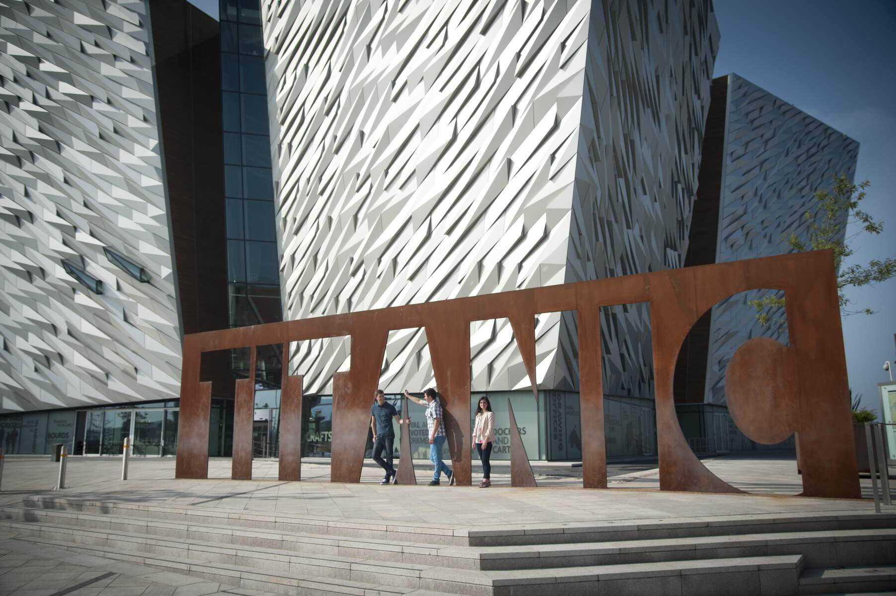 Tre persone camminano davanti al segno di metallo del Titanic fuori dal centro visitatori di Titanic Belfast, con la sua struttura angolare rivestita di metallo.