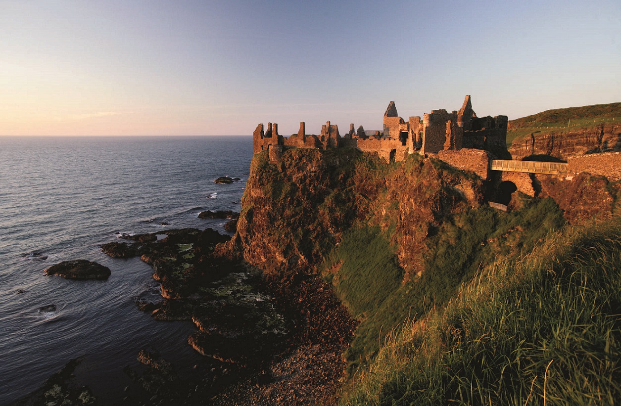 Dunluce Castle ruins cling to a sheer cliffside above crashing waves, glowing gold in late afternoon sun.
