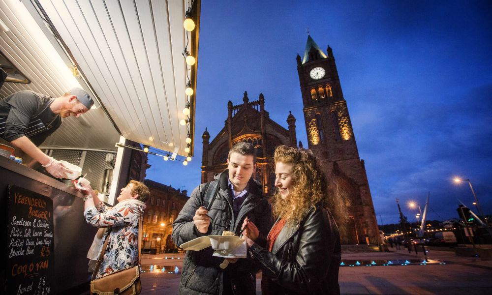 Die Menschen bestellen und essen an einem Imbisswagen vor der beleuchteten Guildhall in der Abenddämmerung in Derry~Londonderry.