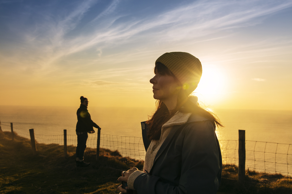 Eine Frau in einer Mütze blickt während des goldenen Sonnenuntergangs auf die See auf der Rathlin-Insel, ein Mann steht weiter entlang des Steigwegs auf dem Kliff.