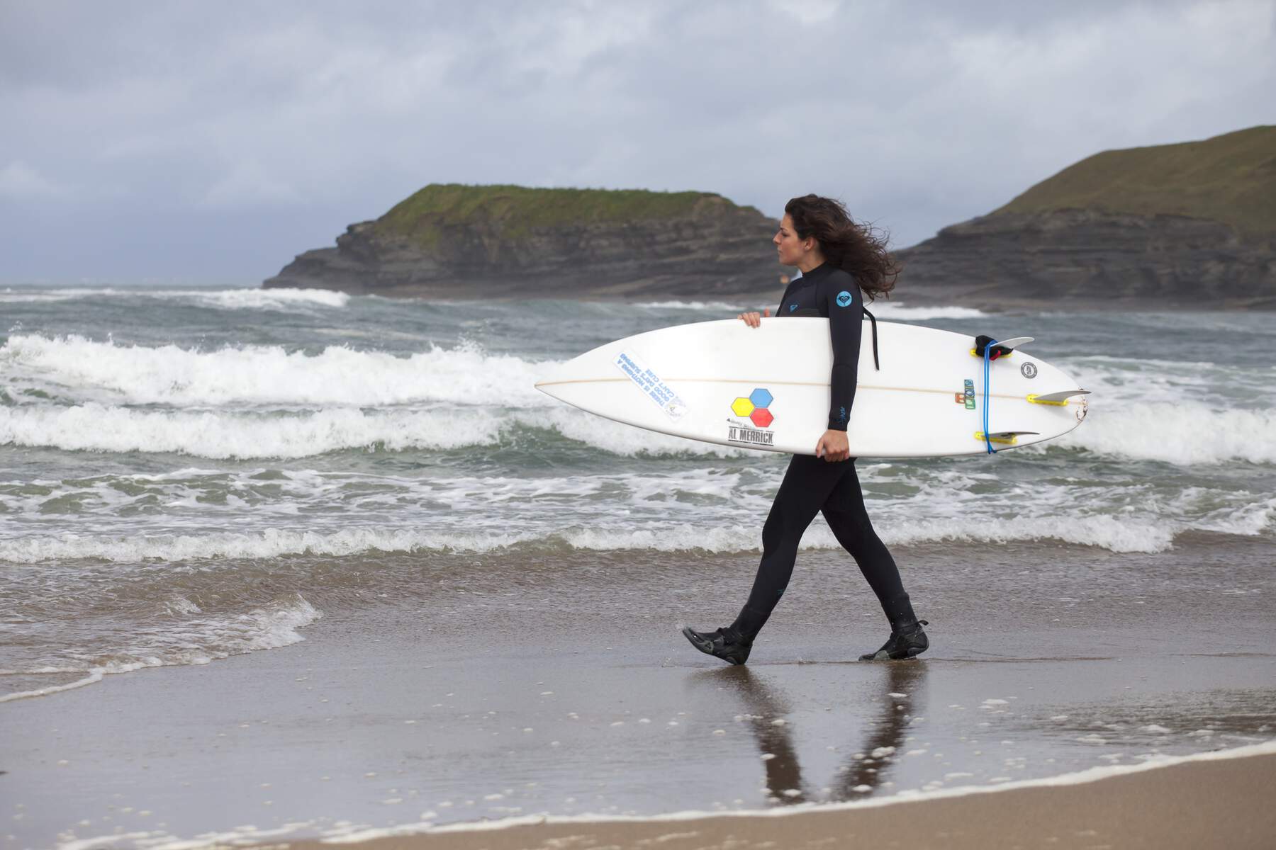 Femme en combinaison de plongée portant une planche de surf le long de la plage de Streedagh, dans le comté de Sligo, avec des vagues et des caps rocheux derrière.