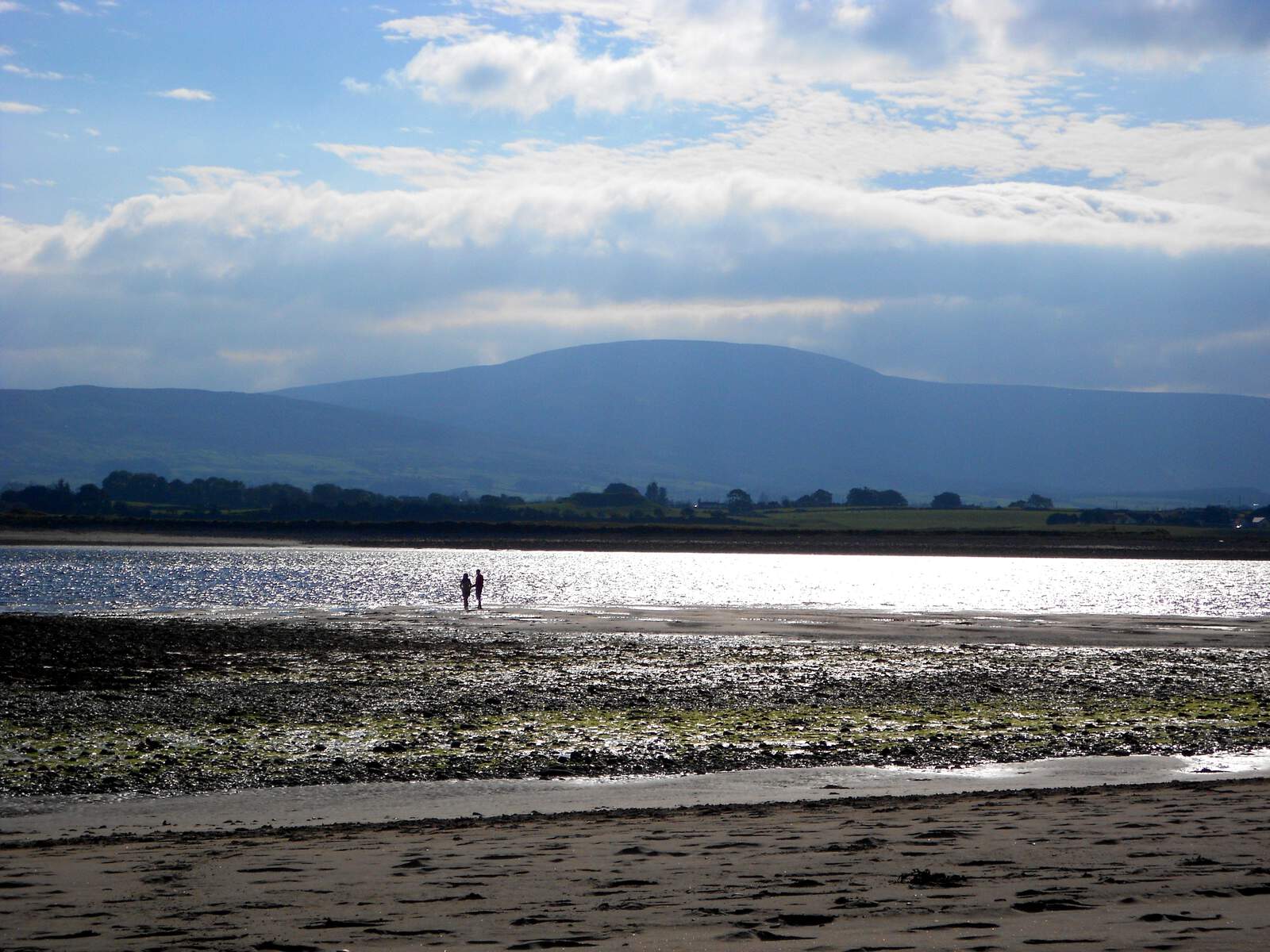 Deux personnes se promènent sur un banc de sable scintillant à marée haute, avec la silhouette de la montagne de Ben Bulben en arrière-plan.
