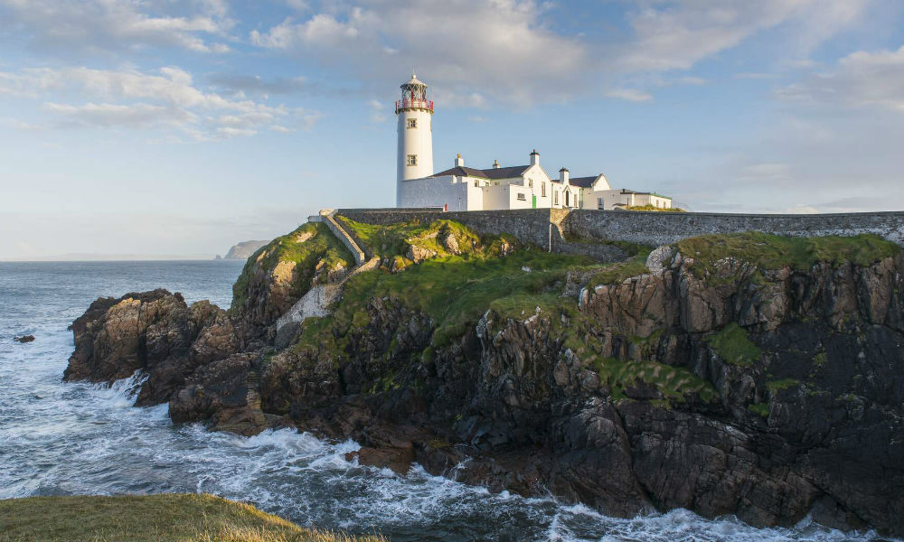 Ein weißer Leuchtturm und das Leuchtturmwärterhaus thronen auf zerklüfteten Klippen an der Fanad-Halbinsel, während die Wellen darunter brechen.