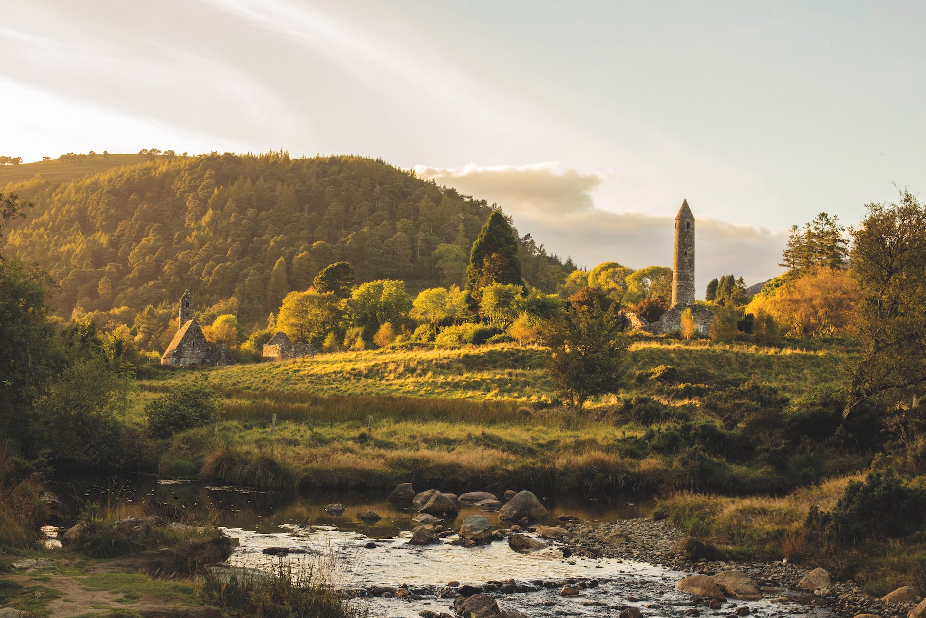 Round tower and monastic ruins surrounded by golden light and woodland in Glendalough, County Wicklow.