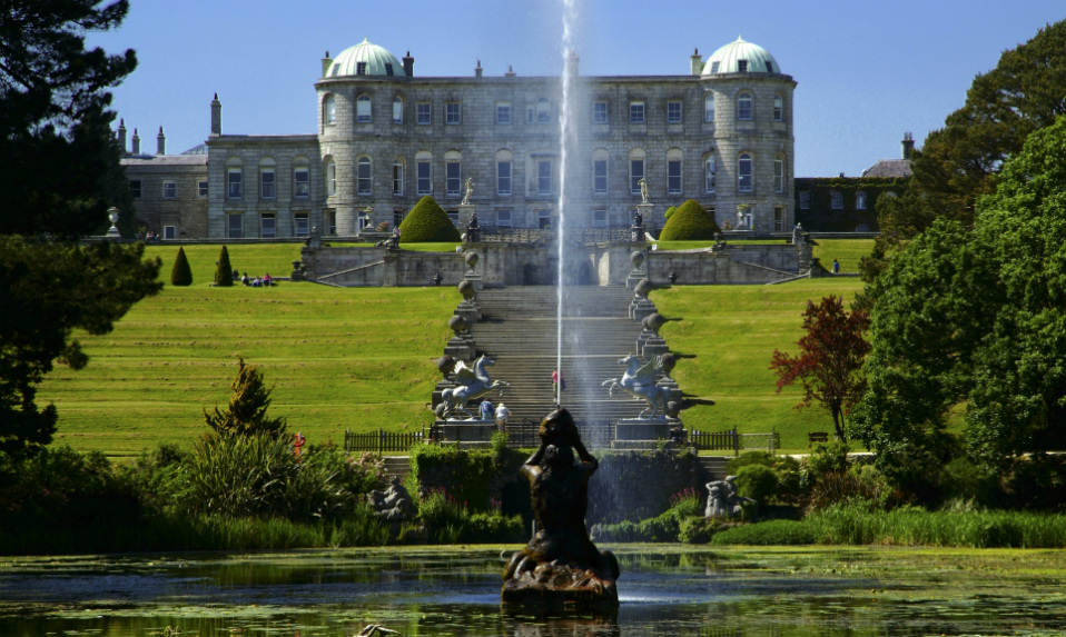 Maison de Powerscourt avec jardins en terrasses, une grande fontaine centrale et des haies sculptées surplombant un étang réfléchissant.