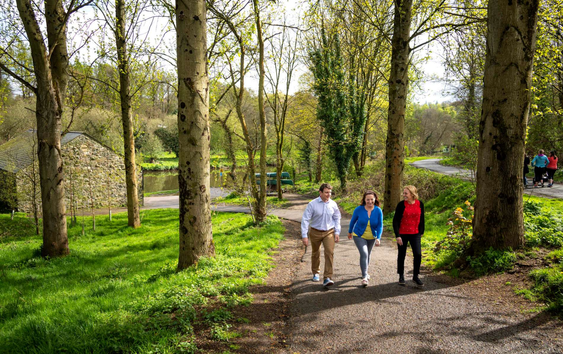 People walking along the River Lagan towpath through trees and past a stone building.