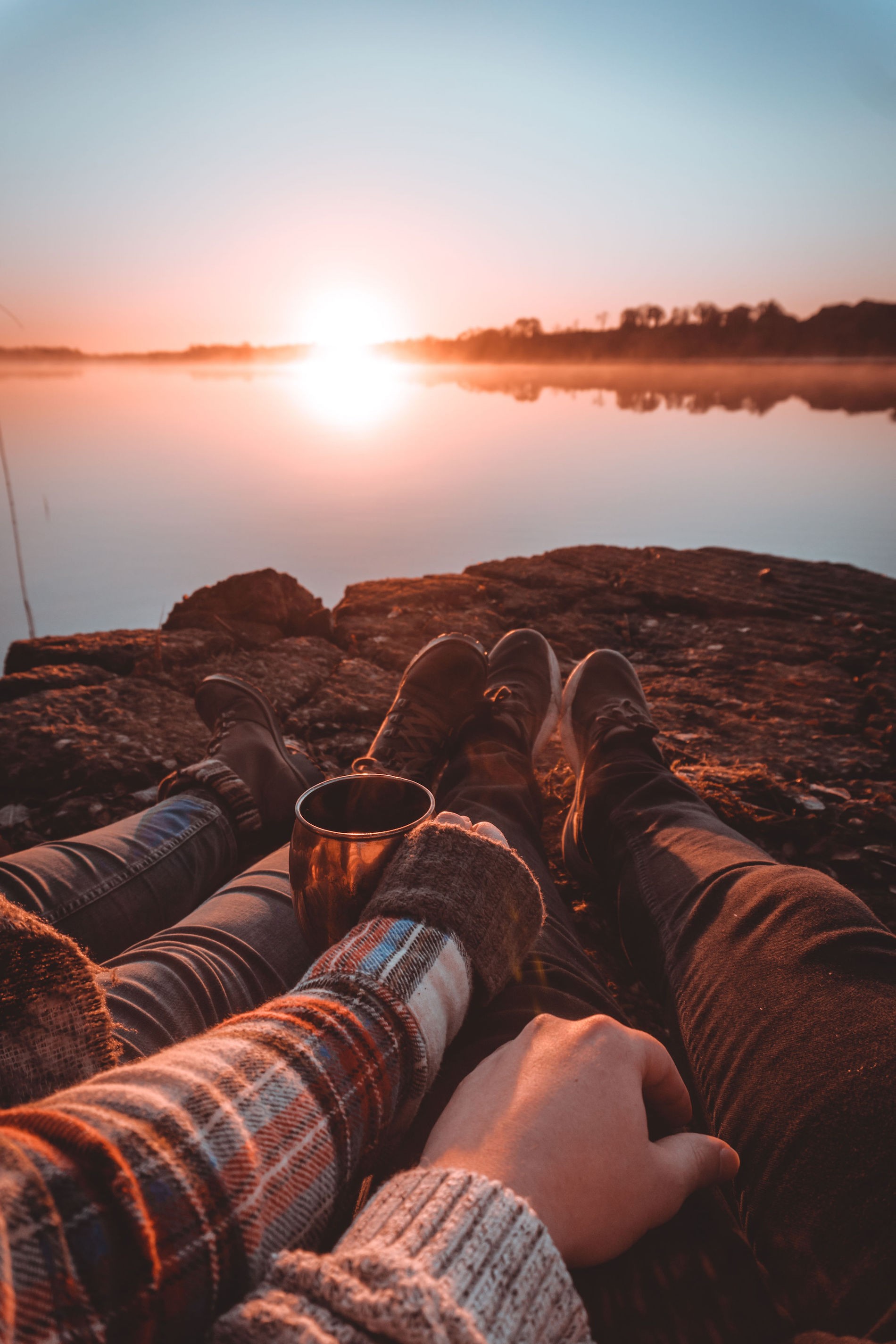 Un couple se détend sur un rivage rocheux en Irlande au lever du soleil avec une boisson chaude, avec vue sur l'eau calme et les arbres lointains.