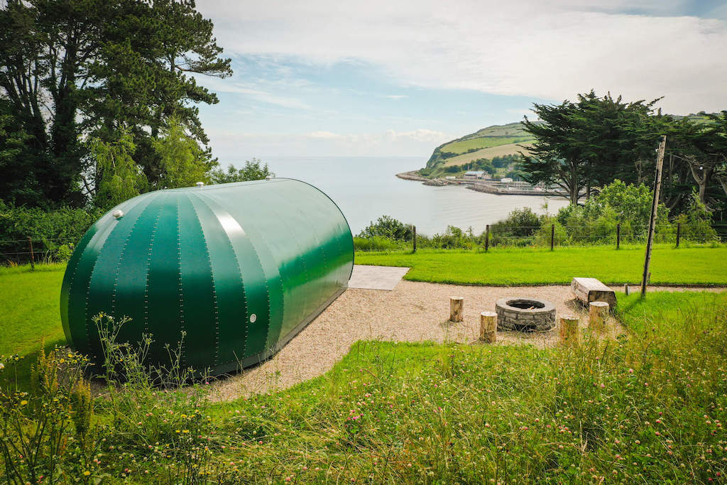 A green glamping pod overlooking the sea and countryside in Glenarm, with firepit, picnic benches, and wildflowers in view.