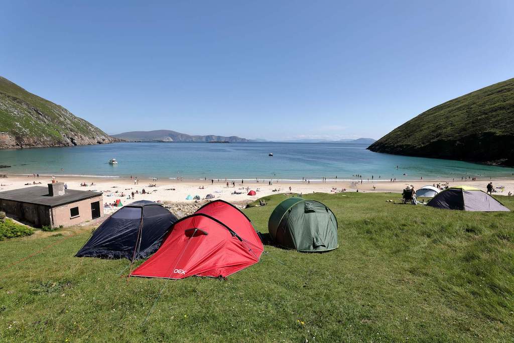 Colourful tents pitched above Keem Bay on Achill Island, with clear blue sea, sunbathers, and green hills in the distance.