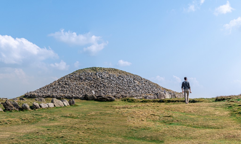 Visitor walking towards a prehistoric stone cairn at Loughcrew, County Meath, under a bright blue sky in rural Ireland.