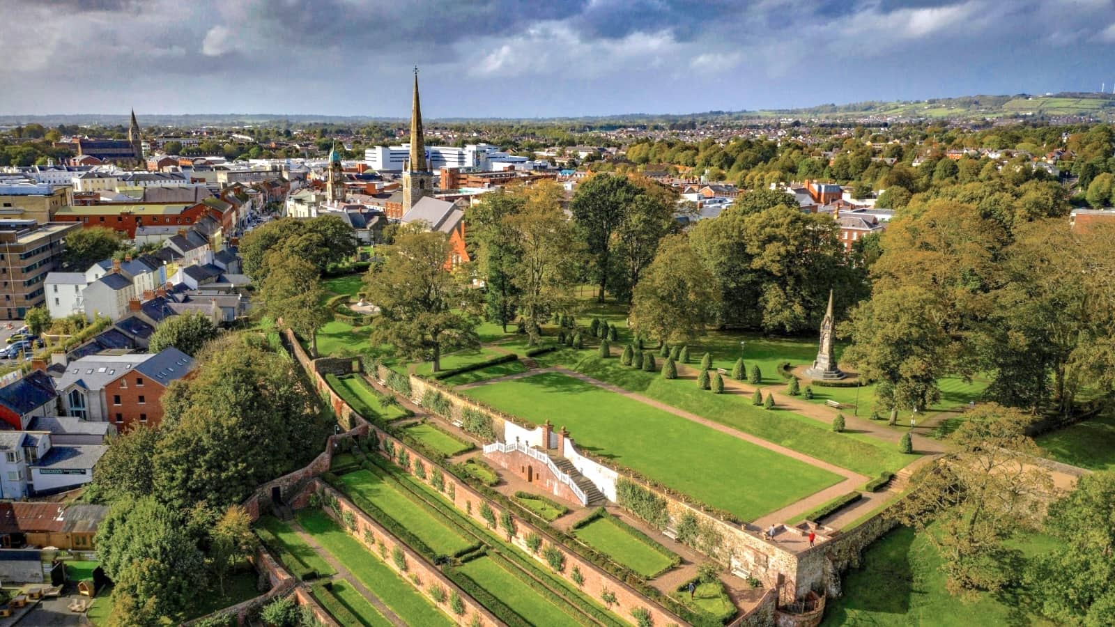 Uitzicht over de stad Lisburn met kasteeltuinen, historische kerken en het omliggende platteland onder een indrukwekkende hemel.