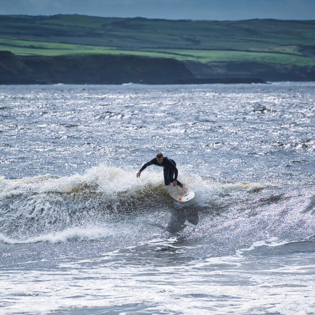 Surfer riding a small breaking wave off the County Clare coast, with rugged green cliffs and shimmering sea in the background.