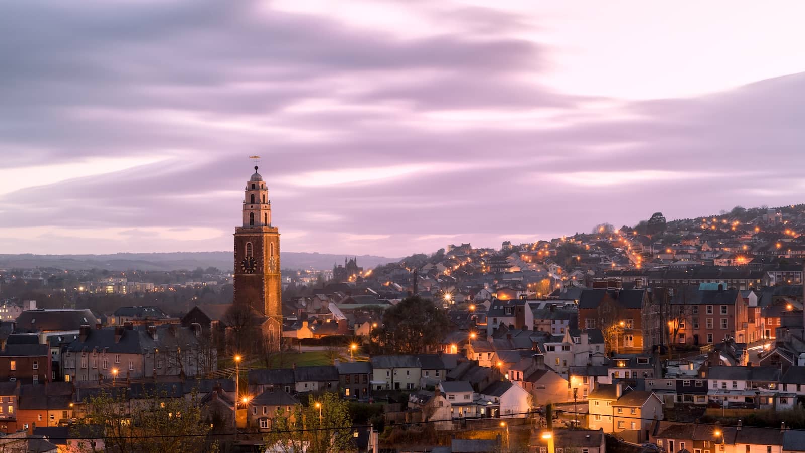Cork City skyline at dusk with St Anne’s Church tower and glowing city lights under evening sky.